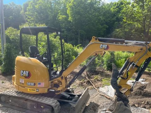 A compact excavator beside a planted tree row.