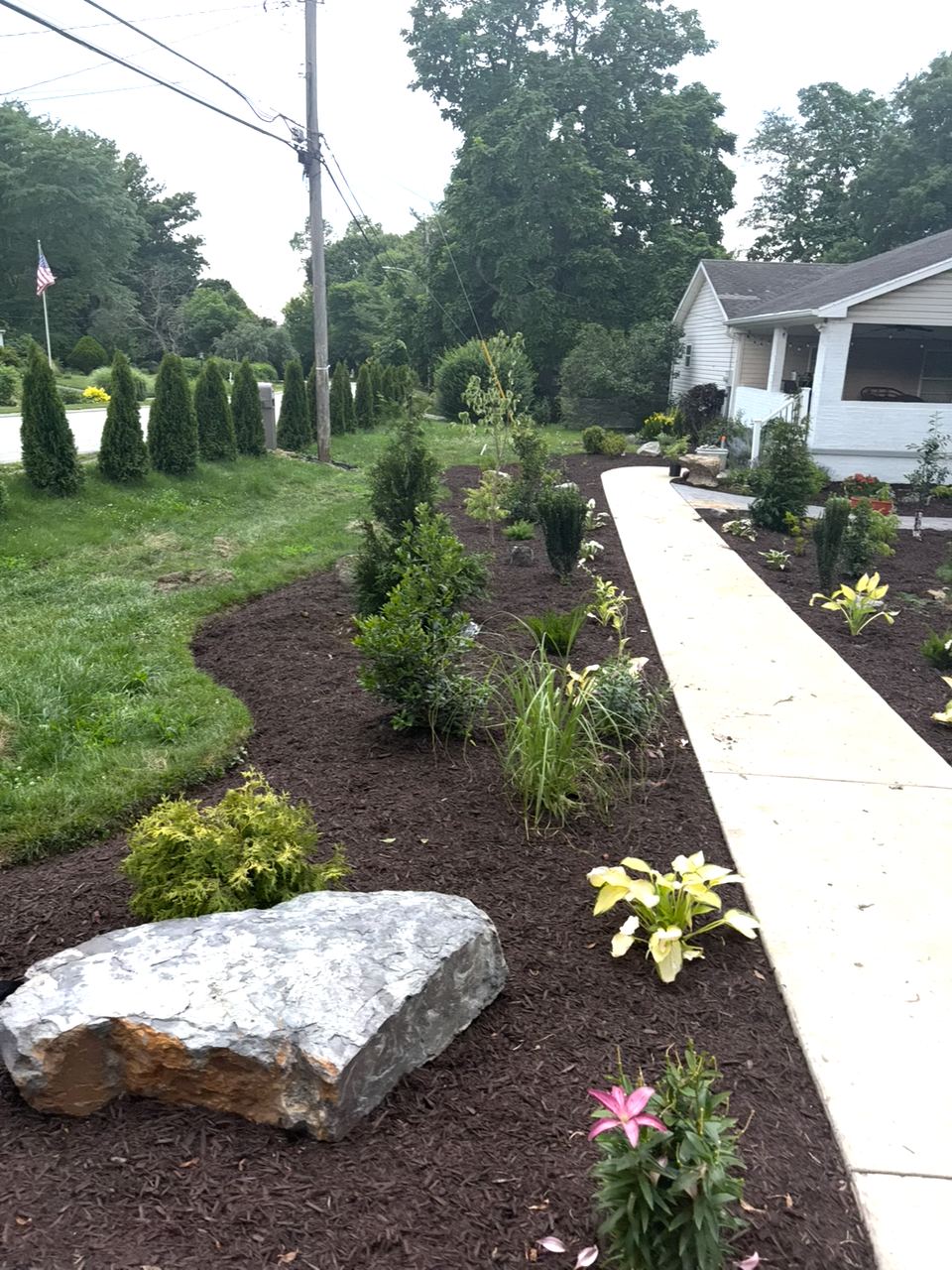 Mixed planting beds and a large rock beside a curved front walkway.