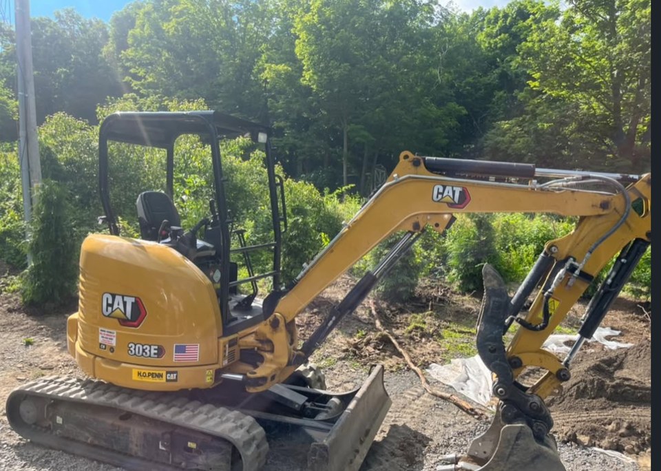 A compact excavator beside a planted tree row.